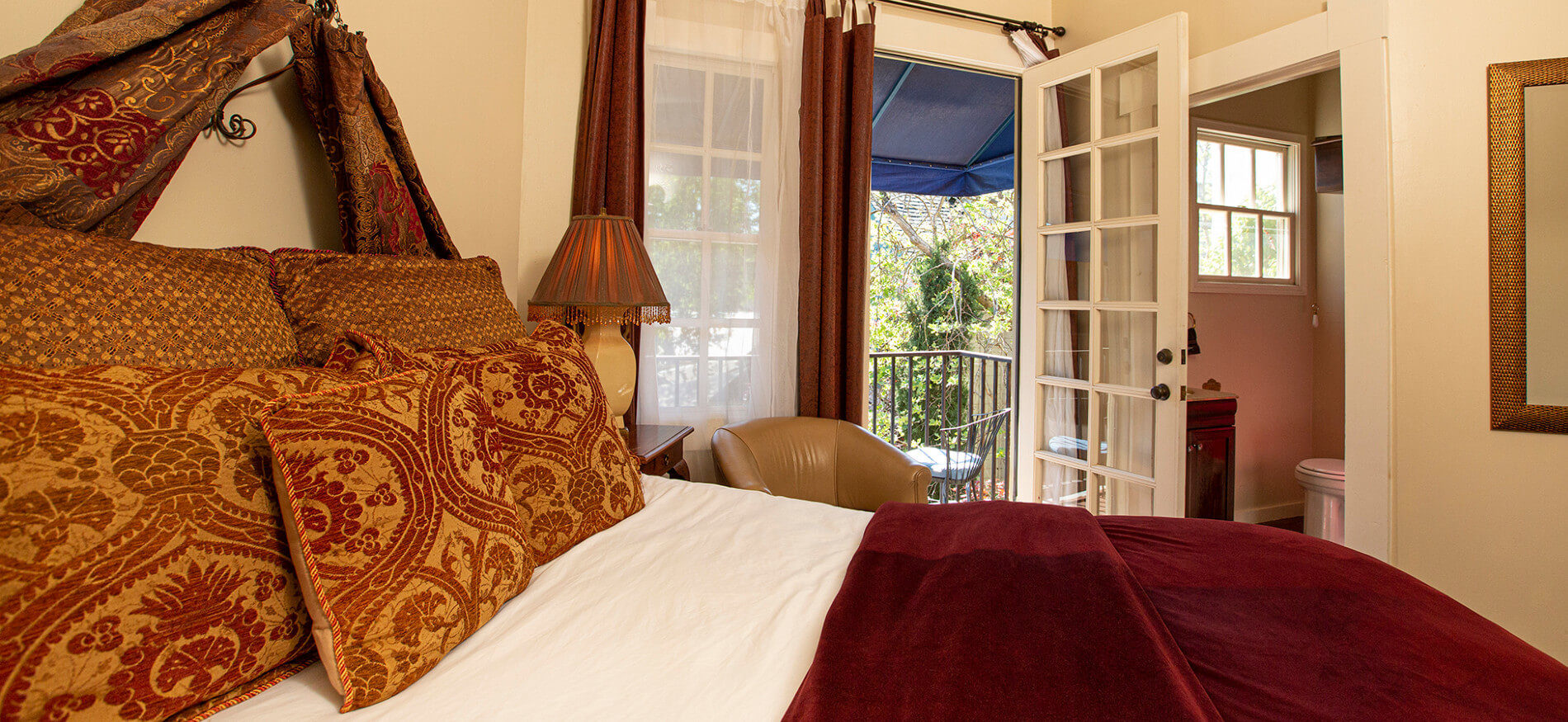 Close up of bed with rich red and gold pillows and wine colored bedspread, bedside lamp, leather chair and French doors