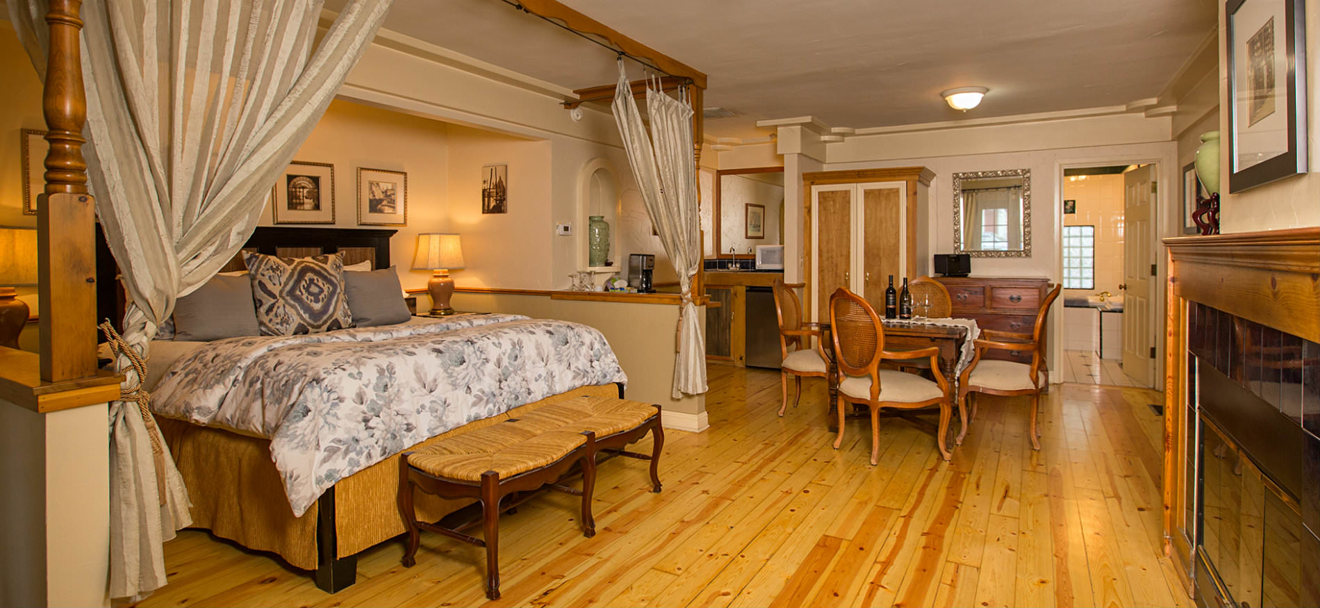 Large room with light wood floors, fireplace, large bed with white and gray floral comforter, and wood table and chairs