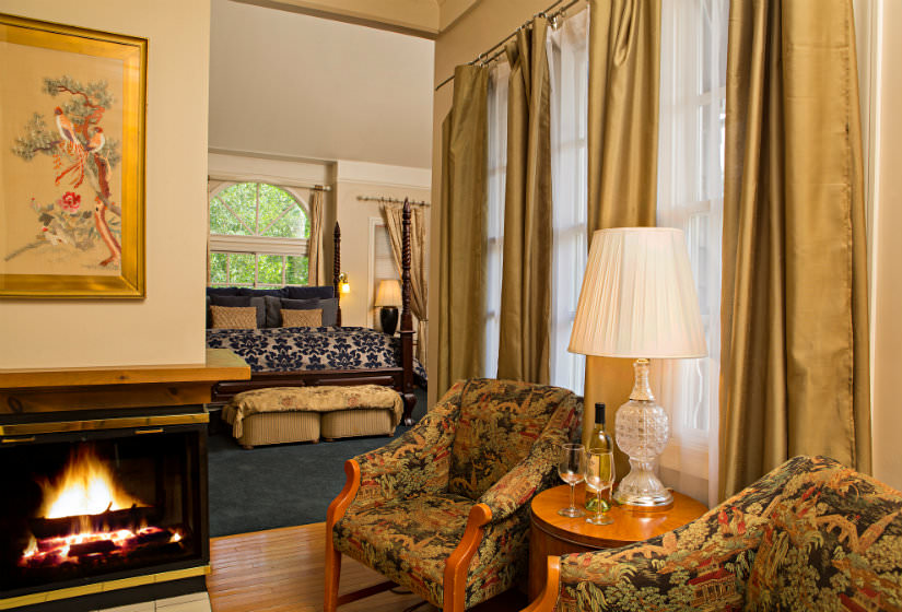 Two chairs with small table on wood floors near fireplace, and blue carpeted bedroom in background with arch top window