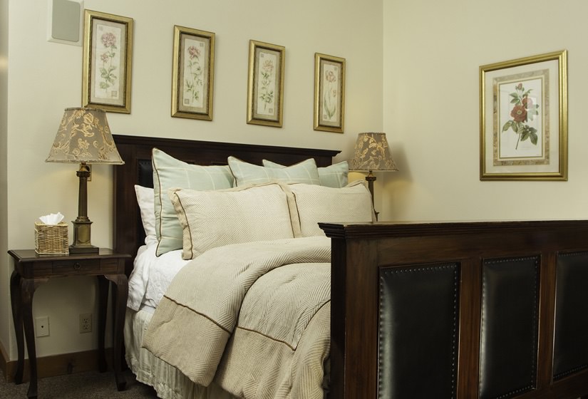 Dark wood and leather bed with ivory comforter, light green and ivory pillows, gold-framed pictures, and nightstands with lamps