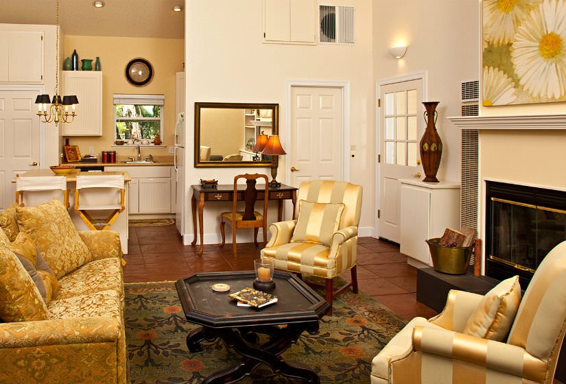 Open area with vaulted ceiling, gold furniture, fireplace, red tile flooring, and kitchen with white cabinets and yellow walls
