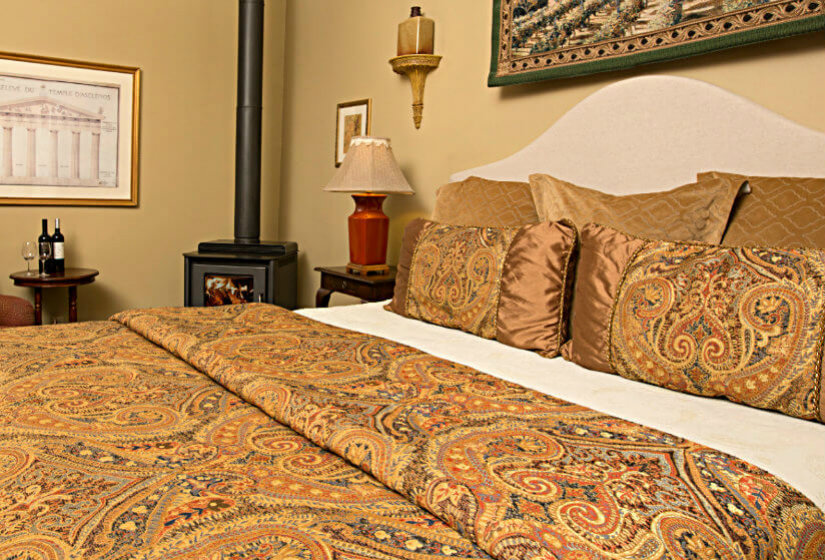 Bed with gold, blue, red, brown paisley pattern comforter, tan walls, red chair with small table, and wood stove