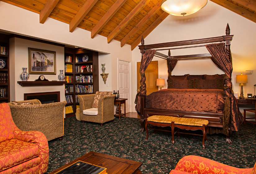 Large vaulted suite with four poster bed, bookshelves flanking fireplace with two chairs, and orange chairs with coffee table