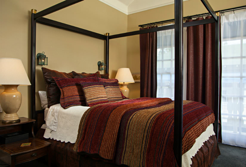 Dark four poster bed with red, brown and gold striped bedspread, large window with red curtains, two nightstands with lamps