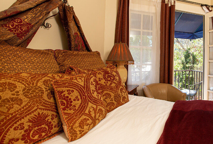 Close up of bed with rich red and gold pillows and wine colored bedspread, bedside lamp, leather chair and French doors
