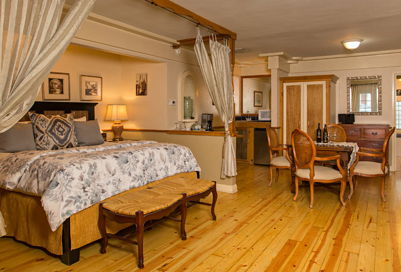 Large room with light wood floors, fireplace, large bed with white and gray floral comforter, and wood table and chairs